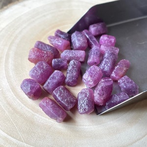 Puede incluir: Una colección de piedras preciosas en bruto de color rosa y morado, probablemente rubíes, en una pala de metal sobre una superficie de madera.
