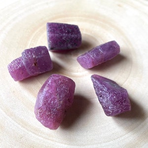 May include: Five rough, purple ruby crystals on a light wood surface.
