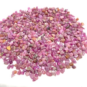 Può includere: Un mucchio di gemme di rubino grezze. Le pietre non tagliate presentano una miscela di tonalità rosa e viola, con alcuni accenti marroni e arancioni. Le pietre variano per dimensioni e forma, creando un aspetto naturale. Sfondo bianco.