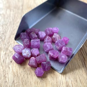 May include: A collection of small, raw, deep pink to magenta-colored ruby gemstones with irregular, faceted shapes. The stones are clustered together inside a metallic silver-toned scoop, displaying natural textures and varying levels of translucency.