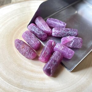 May include: A collection of ten rough, purple ruby gemstones. The gemstones are various sizes and shapes, and are arranged on a wooden surface.
