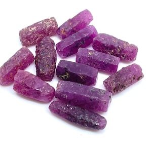 Puede incluir: Un conjunto de cristales de rubí en bruto, cada uno con un tono magenta intenso y una superficie rugosa y texturizada. Las piedras preciosas están dispuestas sobre un fondo blanco, mostrando su estado natural y sin pulir. Ideal para joyería o coleccionismo.