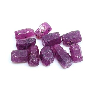 May include: A collection of raw, uncut ruby gemstones. The gemstones are a deep, rich pinkish-red color with a slightly rough texture. The rubies vary in size and shape, with some appearing more elongated and others more blocky.