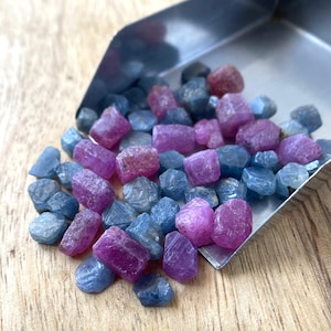 May include: A metal scoop filled with a collection of small, raw, unpolished gemstones in various shades of blue and vibrant pink. The stones have irregular, natural shapes and textures, displaying a mix of translucent and opaque surfaces.