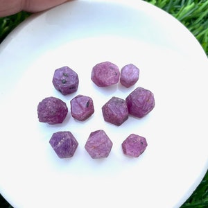 Könnte beinhalten: Eine Sammlung von neun rosafarbenen Rubingesteinen, jeder mit einer facettierten, sechseckigen Form. Die Edelsteine sind auf einem weißen Teller angeordnet.