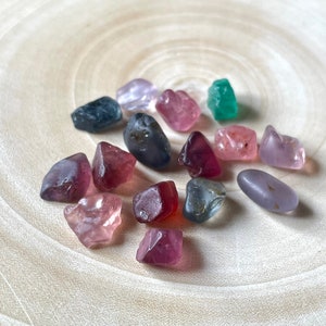 Puede incluir: Una colecci&oacute;n de 15 peque&ntilde;as piedras preciosas facetadas en varios colores, incluyendo rosa, morado, azul, verde y rojo. Las piedras preciosas est&aacute;n dispuestas sobre una superficie de madera clara.