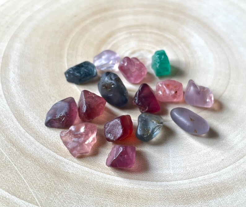 Puede incluir: Una colecci&oacute;n de 15 peque&ntilde;as piedras preciosas facetadas en varios colores, incluyendo rosa, morado, azul, verde y rojo. Las piedras preciosas est&aacute;n dispuestas sobre una superficie de madera clara.