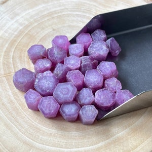 May include: A pile of pink ruby gemstones, with a hexagonal shape, are displayed on a wooden surface. The gemstones are arranged in a loose pile, with some of them overlapping.