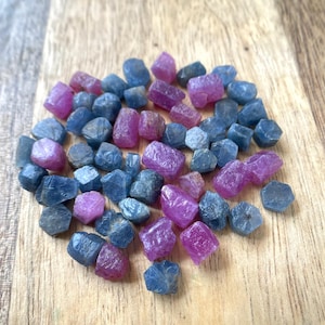 Puede incluir: Una colección de pequeñas piedras preciosas en bruto y sin pulir en tonos azul profundo y rosa vibrante. Estas piezas minerales sueltas e irregulares varían en tamaño y forma, mostrando texturas y facetas naturales en sus superficies.