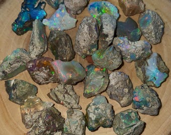 May include: A collection of raw, unpolished opal stones featuring irregular shapes and rough, earthy textures. Each stone displays vibrant flashes of iridescent color, including blue, green, orange, and red, embedded within grey and brown rock.