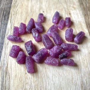 May include: A collection of raw, uncut ruby gemstones in varying sizes and shapes. The gemstones display a deep, rich magenta color and are scattered on a light-colored wooden surface. The rough texture of the stones is visible, highlighting their natural state.