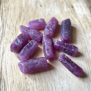 Puede incluir: Una colección de diez piedras preciosas de rubí en bruto de color púrpura. Las piedras preciosas son de diferentes tamaños y formas, pero todas son de un color púrpura intenso.
