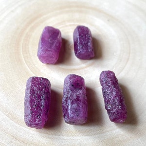 Peut inclure: Cinq cristaux de rubis bruts de couleur violette sur une surface en bois. Les cristaux sont de tailles et de formes différentes, mais ils sont tous d'une couleur violette profonde.