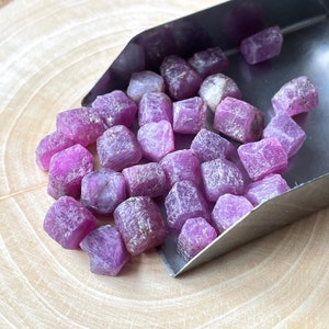 Puede incluir: Una colección de gemas de rubí en bruto sin cortar. Los rubíes son de un color rojo-rosado vibrante, con superficies rugosas y texturizadas. Se muestran en una pequeña cuchara de metal, sobre un fondo de vetas de madera clara.