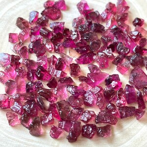 May include: A collection of pink and purple rough tourmaline gemstones. The gemstones are various sizes and shapes, and have a natural, unpolished finish.