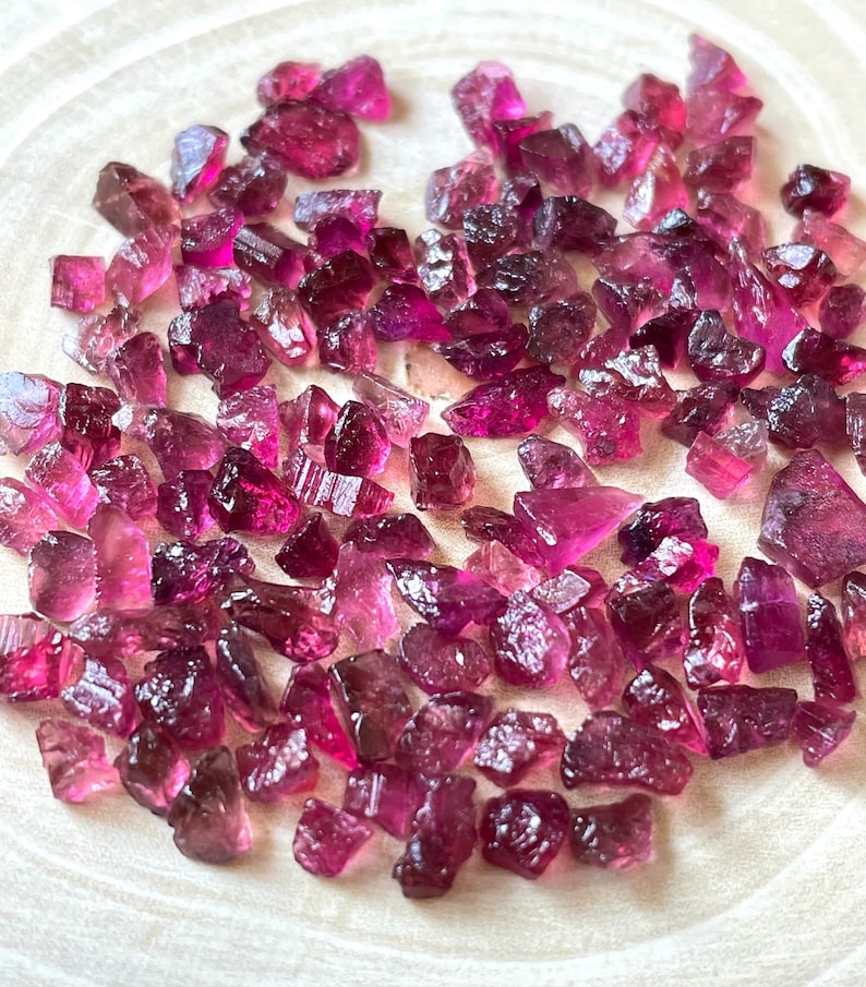 May include: A collection of pink and purple rough tourmaline gemstones. The gemstones are various sizes and shapes, and have a natural, unpolished finish.