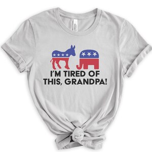 Puede incluir: Una camiseta gris claro con un burro azul y un elefante rojo. El texto "I'M TIRED OF THIS, GRANDPA!" está impreso debajo del gráfico.