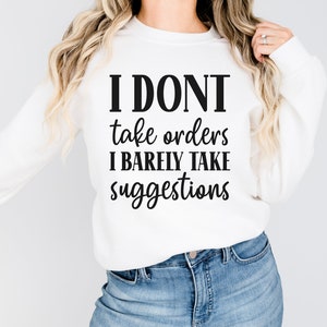 May include: White sweatshirt with the text "I DON'T take orders I BARELY TAKE suggestions" in black. The sweatshirt is long-sleeved and has a crew neck. The model is wearing blue jeans.