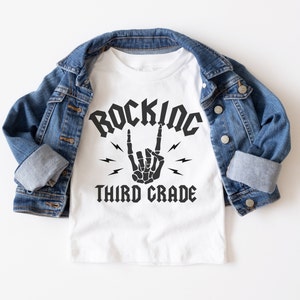 May include: White t-shirt with a black graphic that says "Rockin' Third Grade" with a skeleton hand making the rock and roll sign.