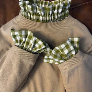 Puede incluir: Un suéter color beige con cuello y puños estampados a cuadros verdes y blancos. El cuello y los puños son con volantes, lo que añade un elemento decorativo al suéter. El suéter está hecho de un material suave.
