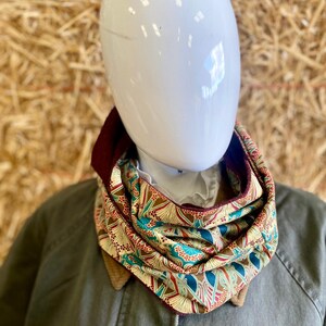 May include: A colorful infinity scarf with a floral and geometric pattern in teal, brown, and red. The scarf is displayed on a mannequin wearing a green jacket. The scarf is approximately 10 inches wide.