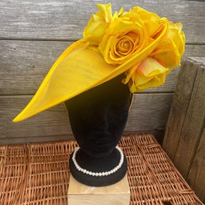 Könnte beinhalten: Ein leuchtend gelber Hut mit breiter Krempe und einer Ansammlung großer gelber Rosen. Der Hut wird auf einem schwarzen Schaufensterkopf mit einer Perlenkette vor einem Holzgrund präsentiert.
