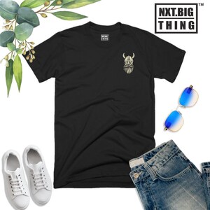 May include: Black t-shirt with a white graphic of a Viking wearing a helmet and a beard with the word "DAD!" underneath. The t-shirt is laid flat on a white background with a pair of white sneakers, blue jeans, and sunglasses.