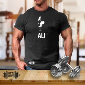 May include: Black t-shirt featuring a white silhouette of Muhammad Ali's face and the word "ALI" printed on the front. The shirt is being worn by a person with muscular arms. Two silver dumbbells are in the foreground.
