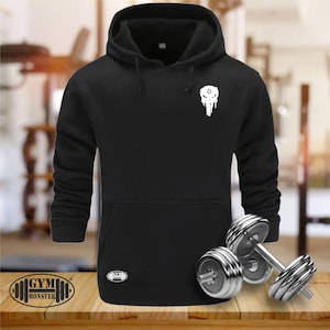 Op de afbeelding: Zwarte hoodie met een witte schedelprint en de tekst "GYM MONSTER" onderaan. De hoodie ligt op een houten oppervlak met twee zilveren dumbbells.