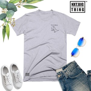 May include: A light gray t-shirt with a graphic of a dinosaur holding a teacup. The text "TEA-REX" is above the dinosaur. The t-shirt has a small logo on the bottom left corner that says "NXT.BIG THING".