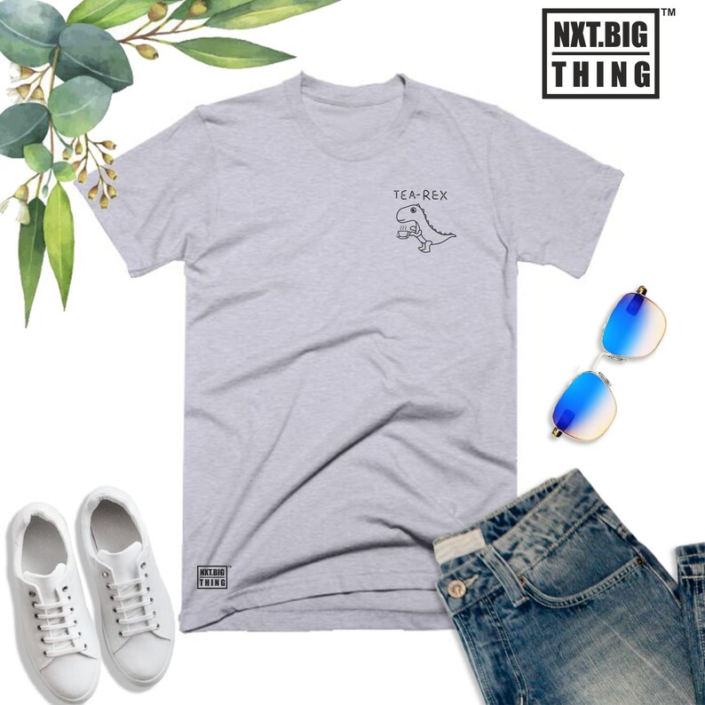 May include: A light gray t-shirt with a graphic of a dinosaur holding a teacup. The text "TEA-REX" is above the dinosaur. The t-shirt has a small logo on the bottom left corner that says "NXT.BIG THING".