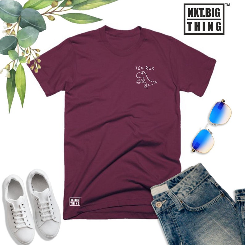 May include: Burgundy t-shirt with a white graphic of a dinosaur holding a cup of tea and the text "Tea-Rex". The shirt is laid out on a white surface with a pair of blue sunglasses, a pair of white sneakers, and a pair of blue jeans.