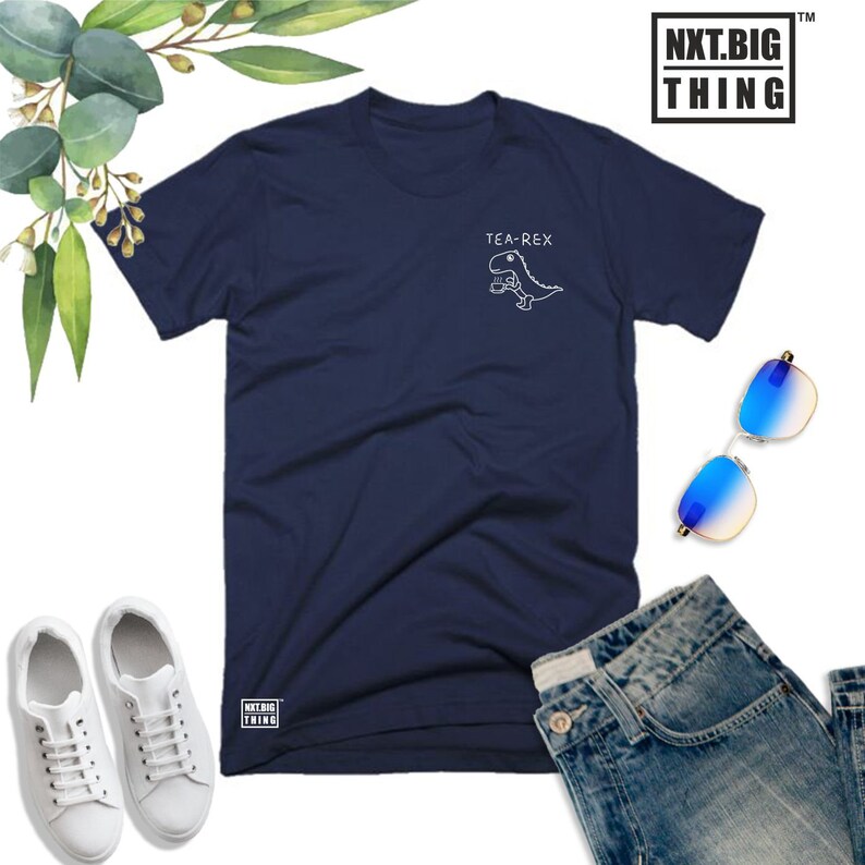 May include: A navy blue t-shirt with a white graphic of a dinosaur holding a cup of tea. The text "TEA-REX" is above the dinosaur. The t-shirt has the text "NXT.BIG THING" in white on the bottom left corner.
