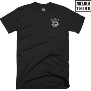 Könnte beinhalten: Schwarzes T-Shirt mit einem kleinen, beigefarbenen Patch auf der linken Brust. Der Patch zeigt ein Design im Vintage-Stil mit dem Text "World's Best Daddy" und einer kleinen Illustration eines Babys.