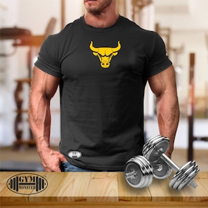 Op de afbeelding: Een zwart T-shirt met een gele stierenkop-afbeelding. Het T-shirt wordt gedragen door een persoon met grote biceps en onderarmen. De tekst "GYM MONSTER" is zichtbaar in de linkerbenedenhoek van de afbeelding.