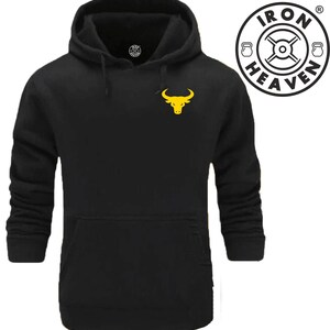Op de afbeelding: Zwarte hoodie met een geel stierenlogo op de voorkant. Het logo is een gestileerde stierenkop met hoorns. De hoodie heeft een kangoeroezak op de voorkant. De tekst "IRON HEAVEN" staat in een cirkel met een halterstang en halters.