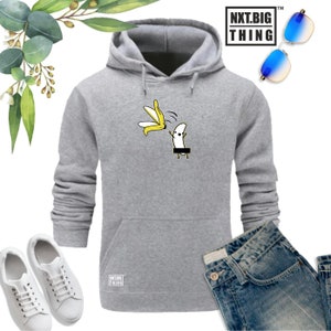 Peut inclure: Un sweat à capuche gris avec une poche sur le devant. Le sweat-shirt présente un motif de banane avec un visage et des bras, tenant une banane pelée. Le texte "NXT.BIG THING" est imprimé sur le sweat-shirt.