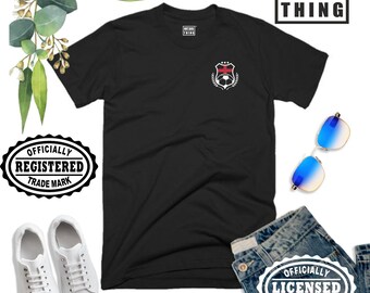 Puede incluir: Camiseta negra con un pequeño diseño de escudo en el pecho, con un balón de fútbol y una corona de laurel. La camiseta se combina con vaqueros azules, zapatillas blancas y gafas de sol. El texto "NXT.BIG THING" es visible.