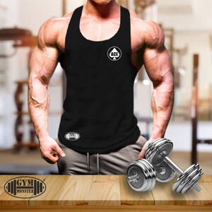Puede incluir: Un tanque negro sin mangas con la palabra "ACE" en un círculo con un símbolo de pica arriba. El tanque también tiene las palabras "GYM MONSTER" en una forma ovalada en la parte inferior. Una persona musculosa lleva el tanque y sostiene pesas.