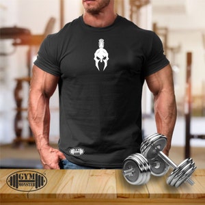 Könnte beinhalten: Schwarzes T-Shirt mit einem weißen Spartanerhelm-Motiv auf der Vorderseite. Das Shirt hat den Text "GYM MONSTER" in der linken unteren Ecke gedruckt. Zwei silberne Hanteln liegen auf einer Holzoberfläche.