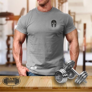 Könnte beinhalten: Ein graues T-Shirt mit einem schwarzen Spartanerhelm-Motiv auf der Vorderseite. Das Shirt wird von einer Person mit muskulösen Armen getragen. Der Text "GYM MONSTER" ist am unteren Rand des Bildes sichtbar.