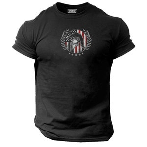 Könnte beinhalten: Schwarzes T-Shirt mit einem Grafikdruck eines spartanischen Helms vor einer amerikanischen Flagge mit einem Lorbeerkranz darum. Der Text "Hero Tribe" befindet sich auf dem Etikett.