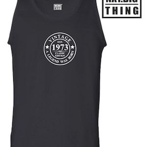 Op de afbeelding: Zwart tanktop met een witte cirkelvormige afbeelding met de tekst "Vintage Est. 1973 Classic Edition A Legend Was Born". De afbeelding bevindt zich op de voorkant van het tanktop. Het tanktop heeft een wit label op de voorkant met de tekst "NXT.BIG THING".