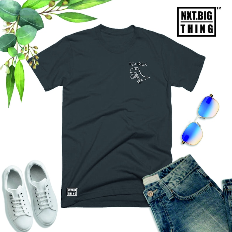 May include: A dark gray t-shirt with a white graphic of a dinosaur holding a teacup. The text "TEA-REX" is above the dinosaur. The shirt has a small logo on the bottom left corner that says "NXT.BIG THING".