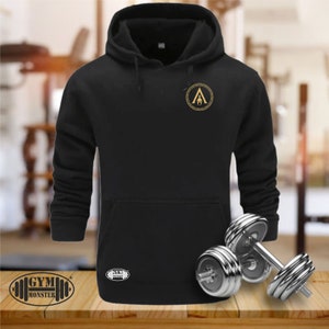 Könnte beinhalten: Schwarzes Kapuzenpullover mit einem goldenen griechischen Krieger-Symbol auf der Vorderseite. Der Pullover liegt auf einer Holzoberfläche mit zwei silbernen Hanteln.