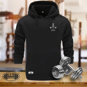 Könnte beinhalten: Schwarzer Kapuzenpullover mit Fronttasche und Kordelzug. Er hat eine weiße Grafik eines Skeletts, das Gewichte hebt, mit dem Wort "DEADLIFT" darunter. Der Pullover hat das "GYM MONSTER"-Logo unten links.