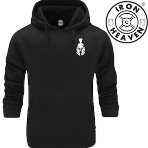 Könnte beinhalten: Schwarzer Kapuzenpullover mit einem weißen Spartanerhelm-Motiv auf der Vorderseite. Der Pullover hat eine Kängurutasche und eine Kapuze mit Kordelzug. Das Iron Heaven Logo befindet sich in der oberen rechten Ecke.