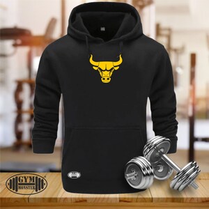 Peut inclure: Sweat-shirt à capuche noir avec un logo de taureau jaune sur le devant. Le sweat-shirt a une poche kangourou et une capuche à cordon. Le texte "GYM MONSTER" est imprimé en bas à gauche de l'image.