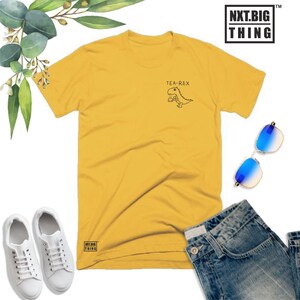 May include: A yellow t-shirt with a black graphic of a dinosaur holding a cup of tea. The text "TEA-REX" is above the dinosaur. The shirt has the text "NXT.BIG THING" on the bottom left corner.