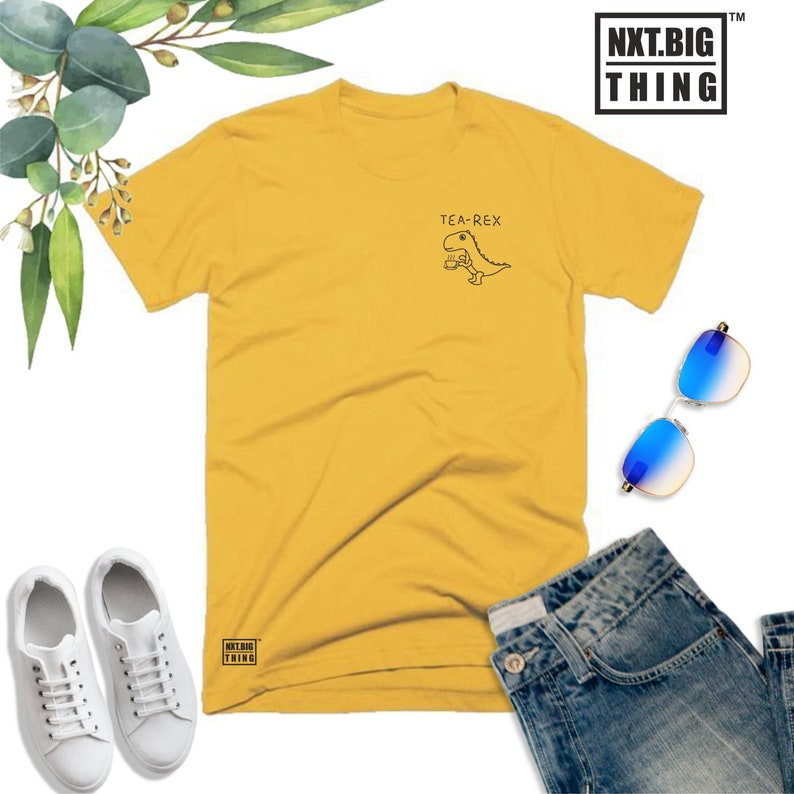 May include: A yellow t-shirt with a black graphic of a dinosaur holding a cup of tea. The text "TEA-REX" is above the dinosaur. The shirt has the text "NXT.BIG THING" on the bottom left corner.